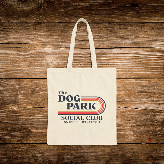 A cream canvas tote bag with 'The Dog Park Social Club' printed on the front, displayed flat against a rustic brown wooden plank background.