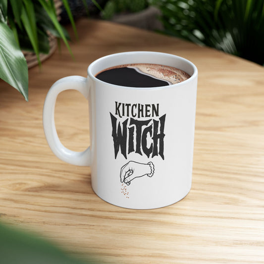 A white ceramic coffee mug with a black graphic that reads 'KITCHEN WITCH' and a small hand sprinkling spices, filled with black coffee and sitting on a light wood table with green plants in the background.