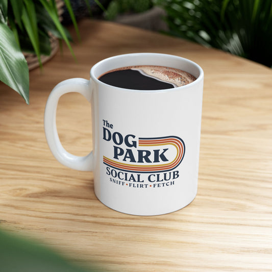 A white ceramic coffee mug filled with black coffee sits on a light wood table. The mug features a retro-style graphic that reads 'The DOG PARK SOCIAL CLUB' with 'SNIFF-FLIRT-FETCH' underneath.