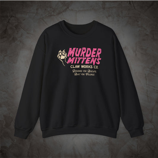 A black crewneck sweatshirt with a graphic on the chest reading 'MURDER MITTENS CLAN WORKS CO' in pink and white, with a fist illustration, displayed on a textured gray background.