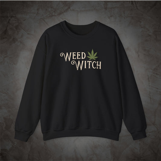 A black crewneck sweatshirt featuring the words 'WEED WITCH' in a distressed cream font with a green cannabis leaf graphic, displayed against a textured dark gray background.