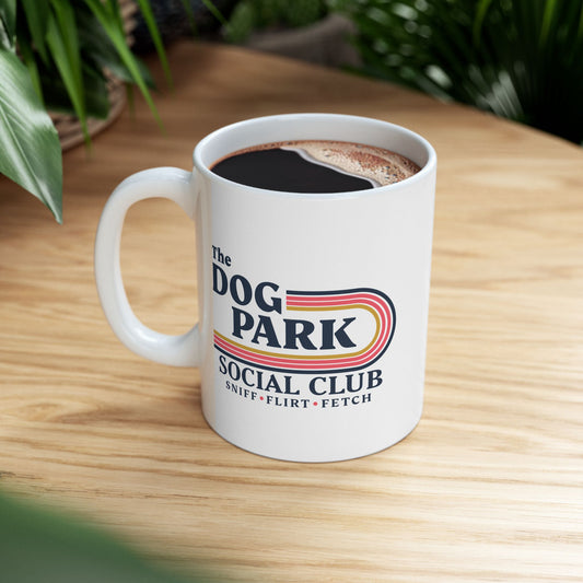A white ceramic coffee mug with a colorful retro-style graphic reading 'The Dog Park Social Club' sits on a light wood table with green plants blurred in the background.