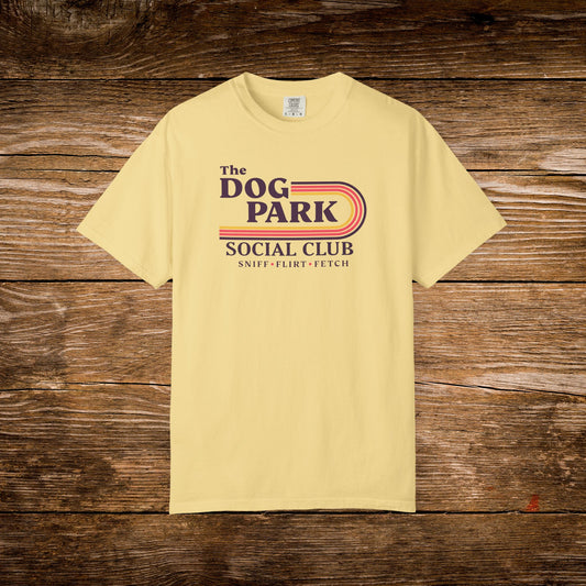 A pale yellow t-shirt with a retro-style graphic reading 'The Dog Park Social Club' and 'Sniff - Flirt - Fetch' in dark blue and orange-red tones, laid flat on a rustic brown wooden background.