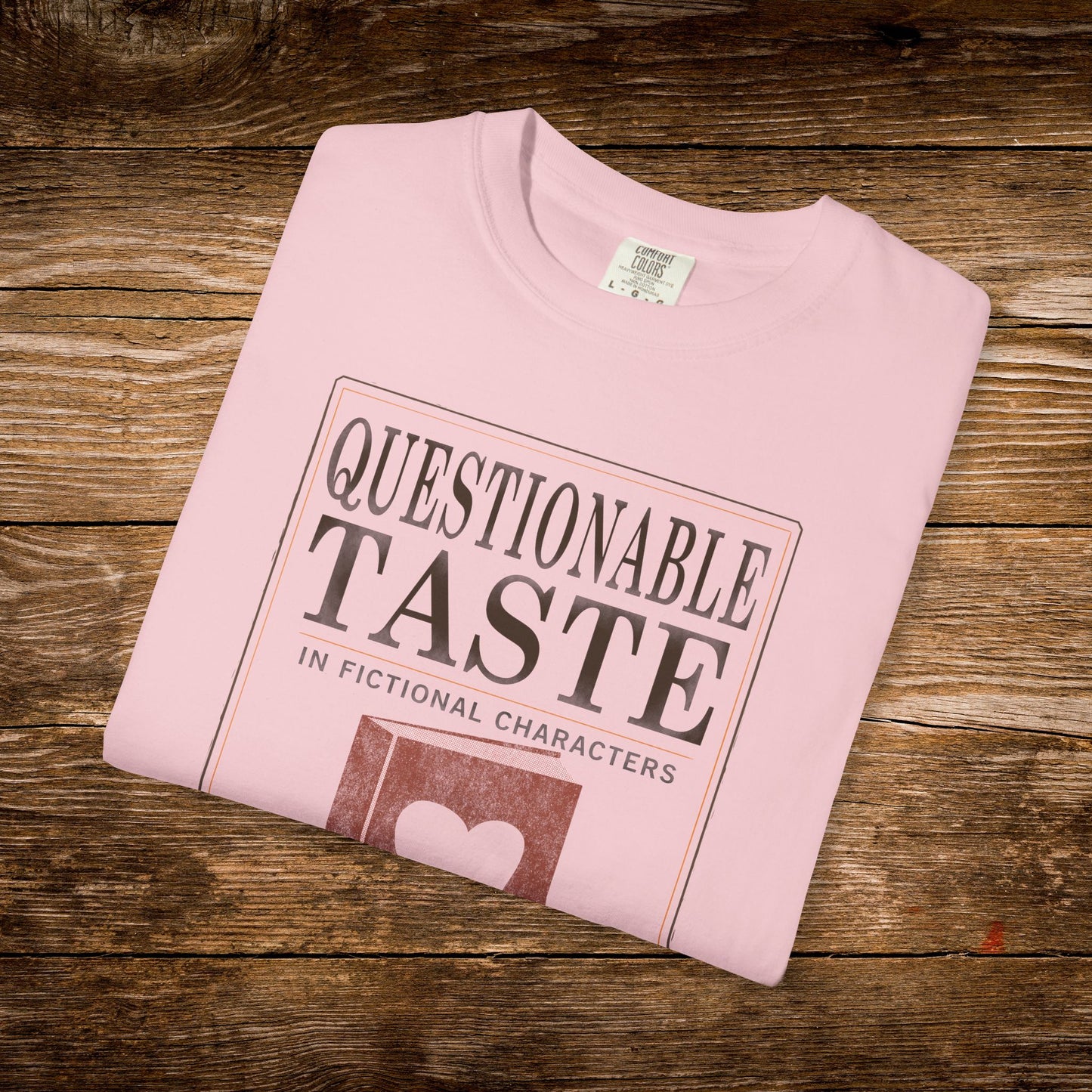 Folded pink t-shirt with 'Questionable Taste' text on a wooden surface
