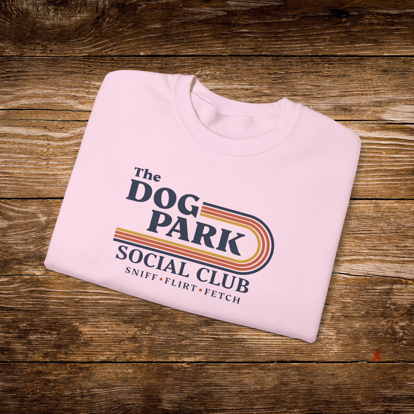 A light pink folded t-shirt with a retro-style graphic reading 'The Dog Park Social Club' and 'Sniff • Flirt • Fetch' in dark purple text with yellow, orange, and red stripes, displayed on a rustic wooden background. The design text reads "Sniff, Flirt, Fetch."