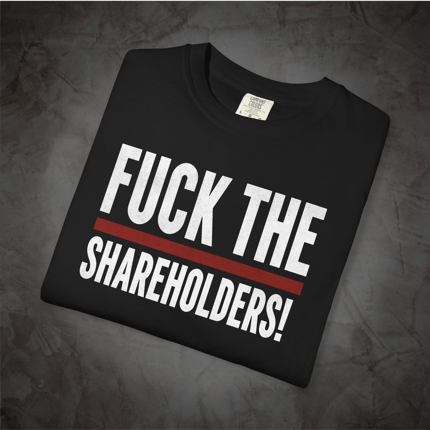 A folded black t-shirt with the text 'FUCK THE SHAREHOLDERS!' printed in bold white letters, separated by a thick red horizontal line, displayed on a textured gray background.