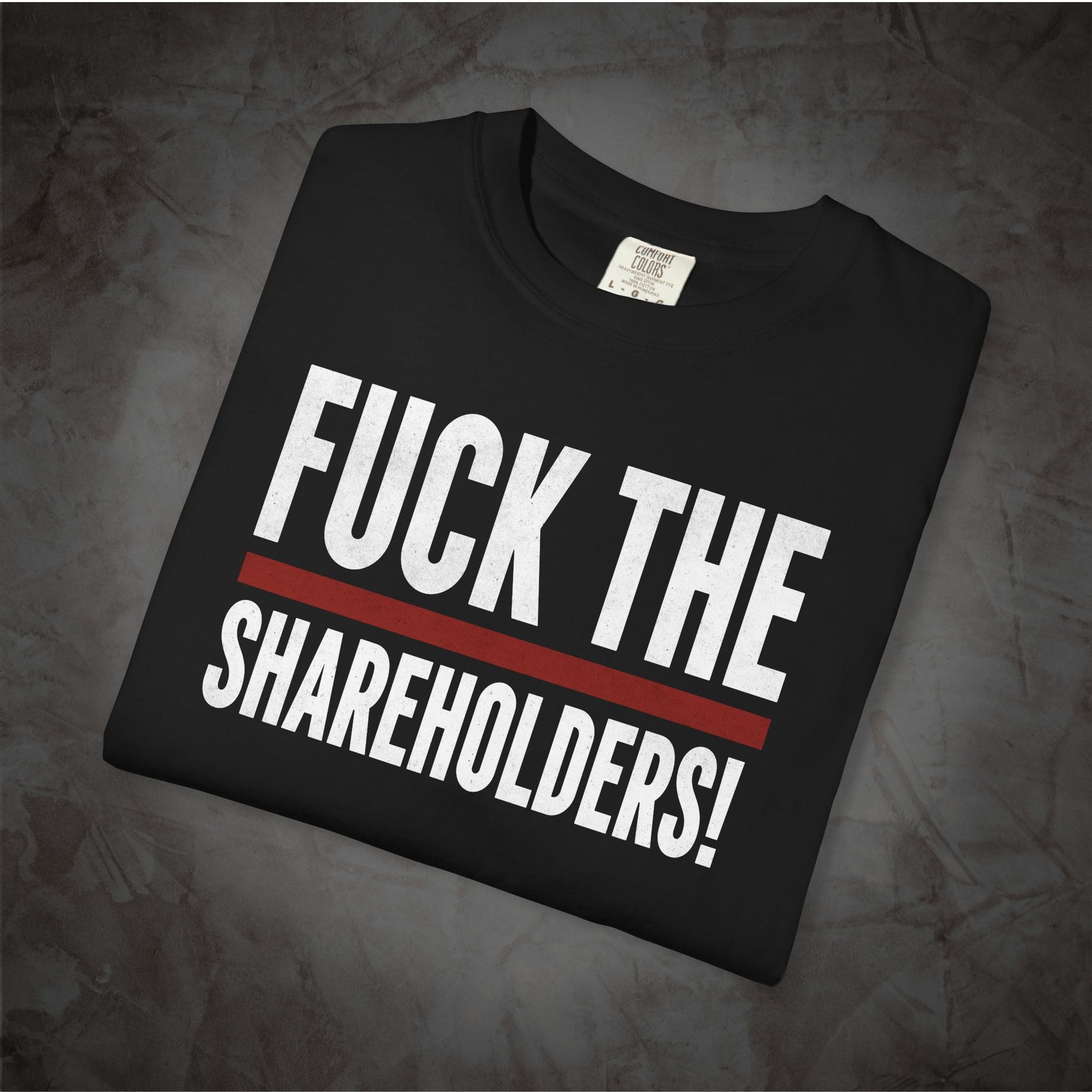 A folded black t-shirt with the text 'FUCK THE SHAREHOLDERS!' printed in bold white letters, separated by a thick red horizontal line, displayed on a textured gray background.