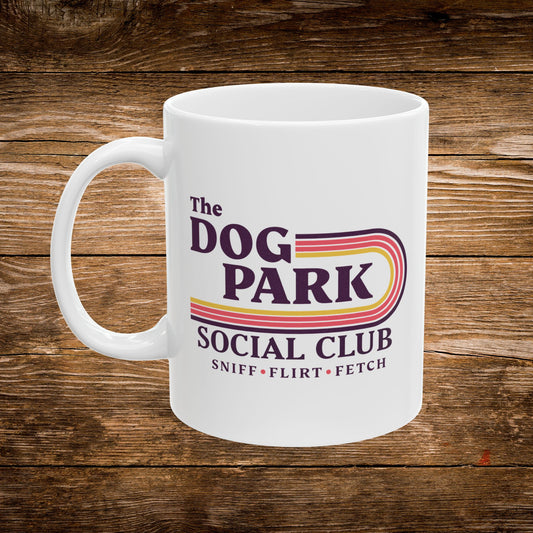 A white ceramic coffee mug with a colorful retro-style graphic that reads 'The DOG PARK SOCIAL CLUB' and 'SNIFF • FLIRT • FETCH' on a rustic brown wooden background.