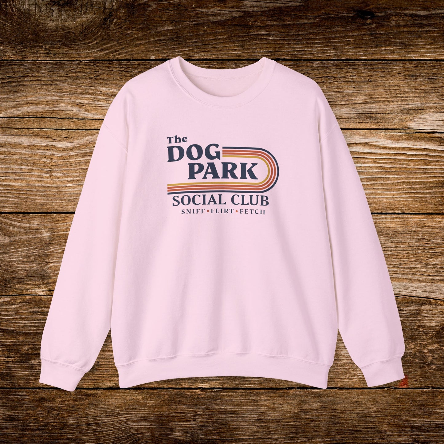A light pink crewneck sweatshirt featuring a retro-style graphic that reads 'The DOG PARK SOCIAL CLUB' with 'SNIFF • FLIRT • FETCH' underneath, displayed flat on a rustic brown wooden plank background. The design text reads "Sniff, Flirt, Fetch."