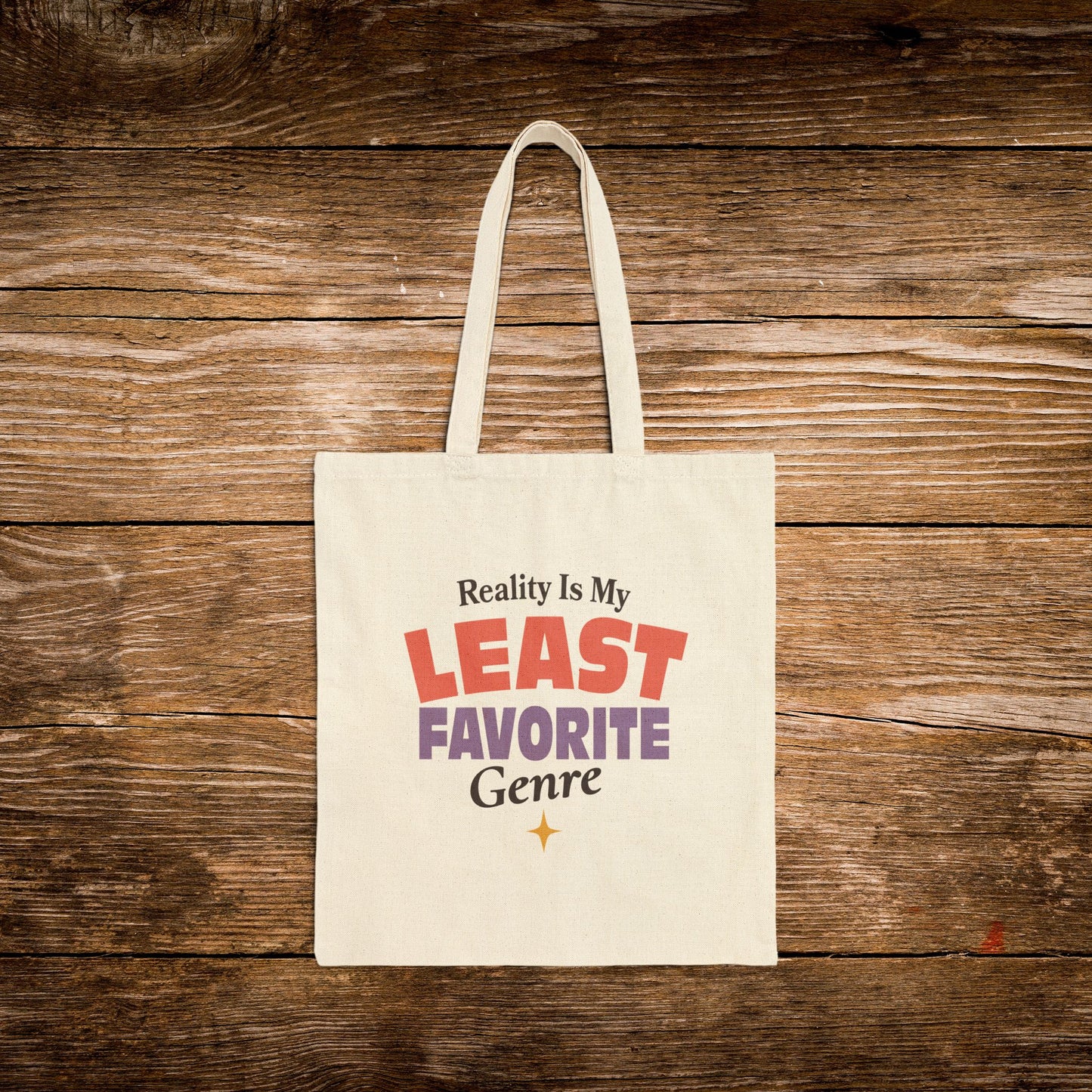 A cream canvas tote bag with the printed phrase 'Reality Is My LEAST FAVORITE Genre' in red, purple, and black lettering, displayed flat against a rustic brown wooden plank background.