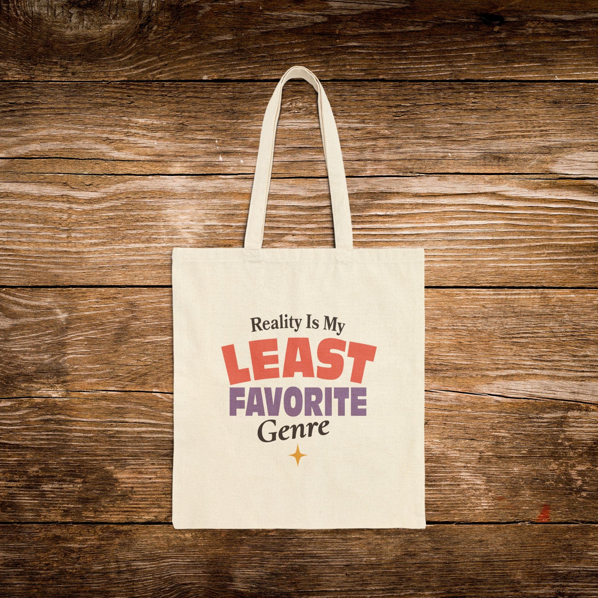A cream canvas tote bag with the printed phrase 'Reality Is My LEAST FAVORITE Genre' in red, purple, and black lettering, displayed flat against a rustic brown wooden plank background.