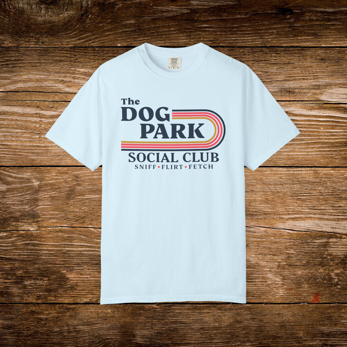 A light blue short-sleeve t-shirt with a retro-style graphic reading 'The Dog Park Social Club' and 'Sniff-Flirt-Fetch' in dark blue text with red and orange stripes, laid flat on a rustic brown wooden background.