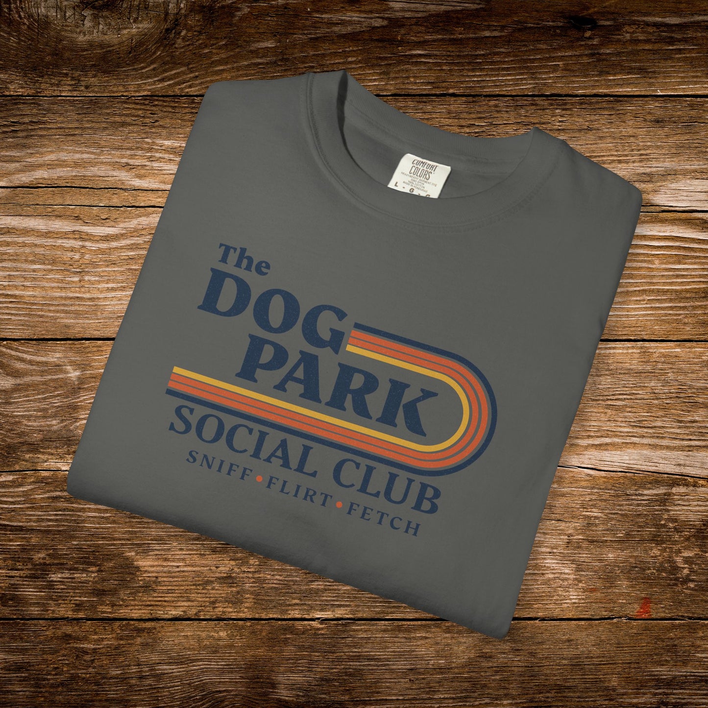 A folded dark gray t-shirt with a retro-style graphic reading 'The Dog Park Social Club' and 'Sniff, Flirt, Fetch' in blue, orange, and yellow, displayed on a rustic wooden background.