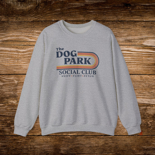 A light heather gray crewneck sweatshirt featuring a retro-style graphic that reads 'The DOG PARK SOCIAL CLUB' with 'SNIFF - FLIRT - FETCH' underneath, displayed flat against a rustic wooden plank background.