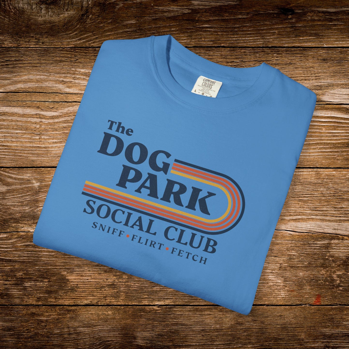 A folded light blue t-shirt with a retro-style graphic reading 'The DOG PARK SOCIAL CLUB' and 'SNIFF * FLIRT * FETCH' in dark blue text with orange, yellow, and red stripes, displayed on a rustic wooden background. The design text reads "Sniff, Flirt, Fetch."