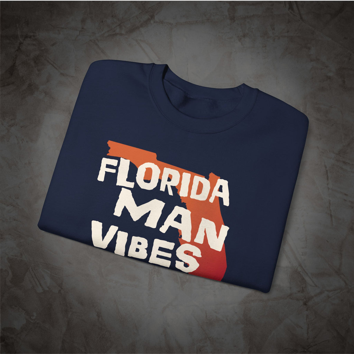 A navy blue folded t-shirt with a graphic print featuring the words 'FLORIDA MAN VIBES' in white over a red-orange silhouette of the state of Florida, displayed on a textured gray background.