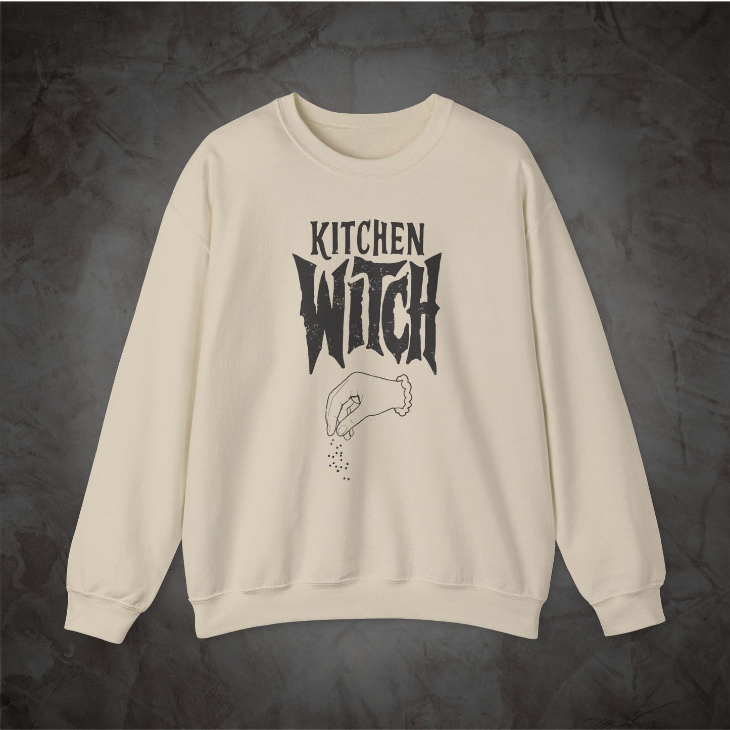 An off-white crewneck sweatshirt featuring a black graphic print that reads 'KITCHEN WITCH' above a hand sprinkling seasoning, displayed flat against a dark textured background.