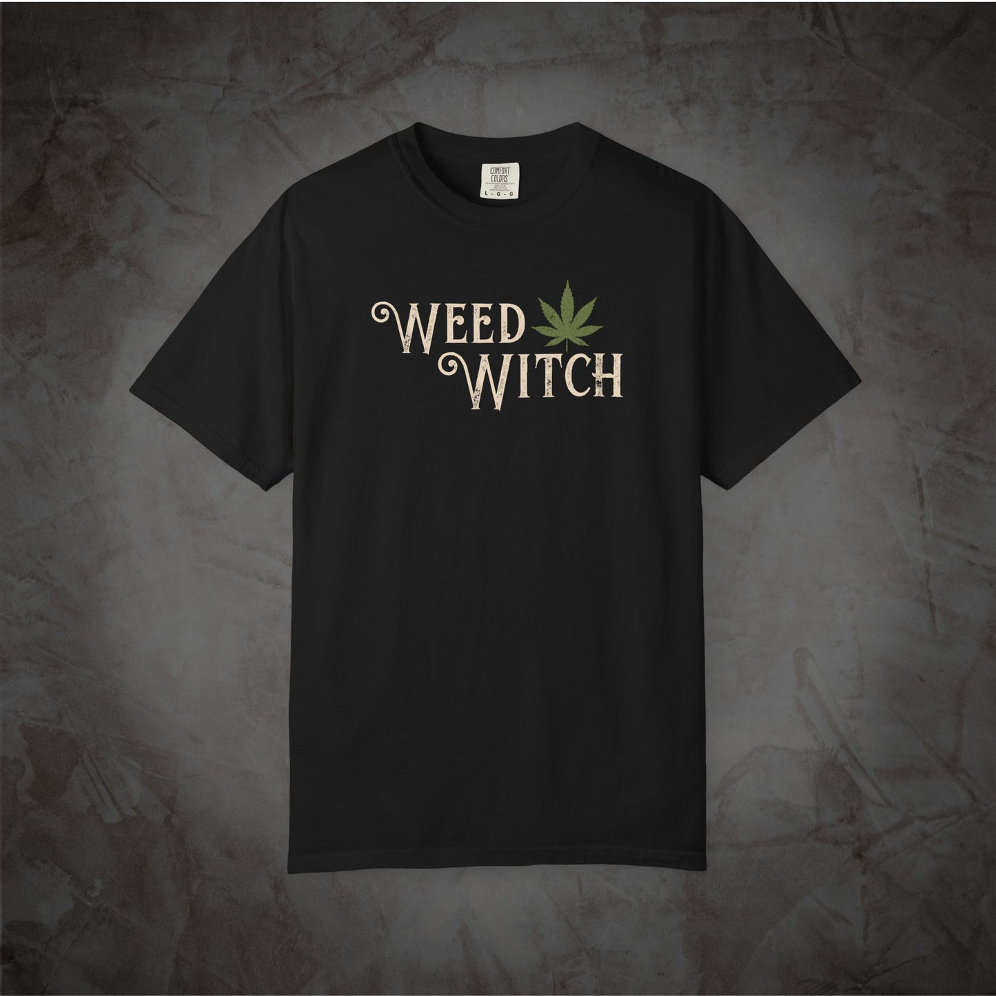 A black short-sleeve t-shirt with a graphic on the chest reading 'WEED WITCH' in a distressed cream font, accompanied by a green cannabis leaf. The shirt is displayed flat against a textured, dark gray background.