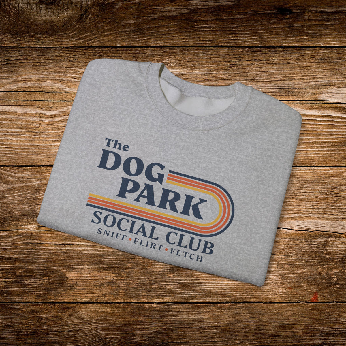 A folded heather gray t-shirt with a retro-style graphic reading 'The Dog Park Social Club' and 'Sniff, Flirt, Fetch' in navy, orange, and yellow, displayed on a rustic wooden background.
