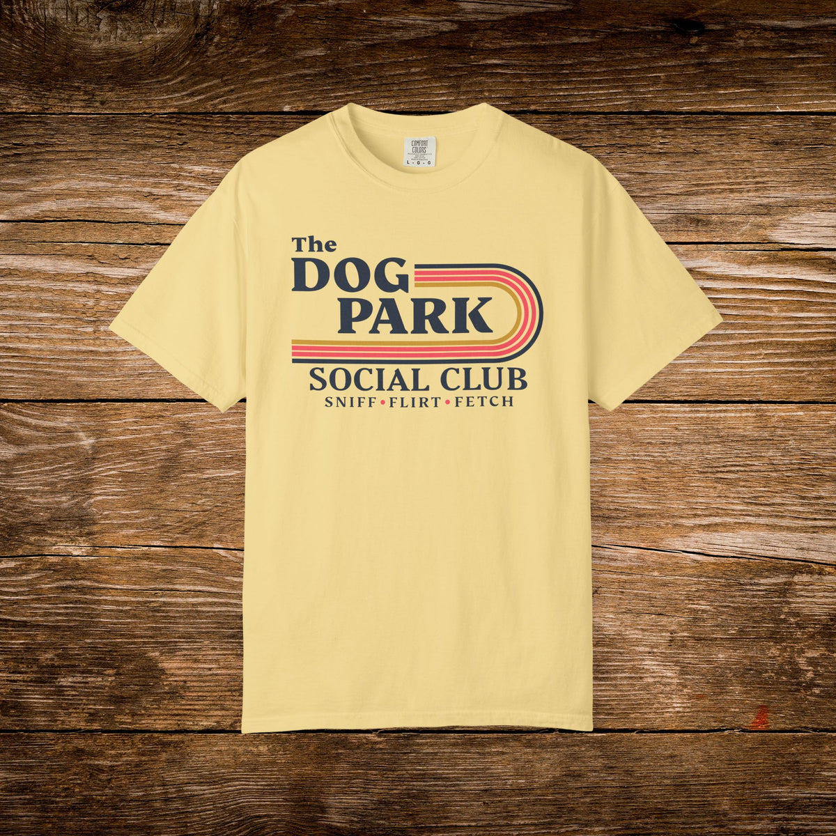 A pale yellow short-sleeve t-shirt with a retro-style graphic reading 'The DOG PARK SOCIAL CLUB' and 'SNIFF - FLIRT - FETCH' laid flat on a rustic dark wood plank background.