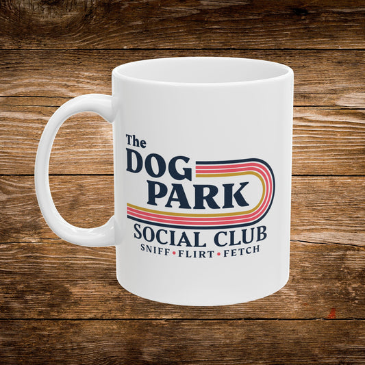 A white ceramic coffee mug with a colorful retro-style graphic that reads 'The DOG PARK SOCIAL CLUB' and 'SNIFF • FLIRT • FETCH' on a rustic brown wood plank background.