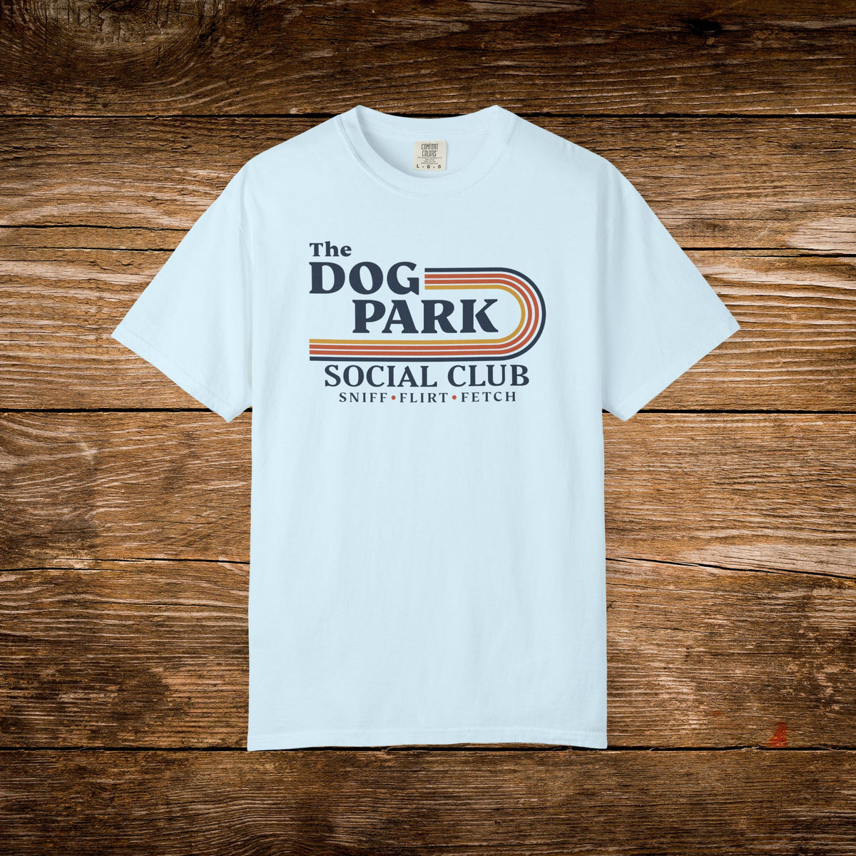 A light blue short-sleeve t-shirt with a retro-style graphic reading 'The DOG PARK SOCIAL CLUB' and 'SNIFF - FLIRT - FETCH' laid flat on a rustic brown wooden background.