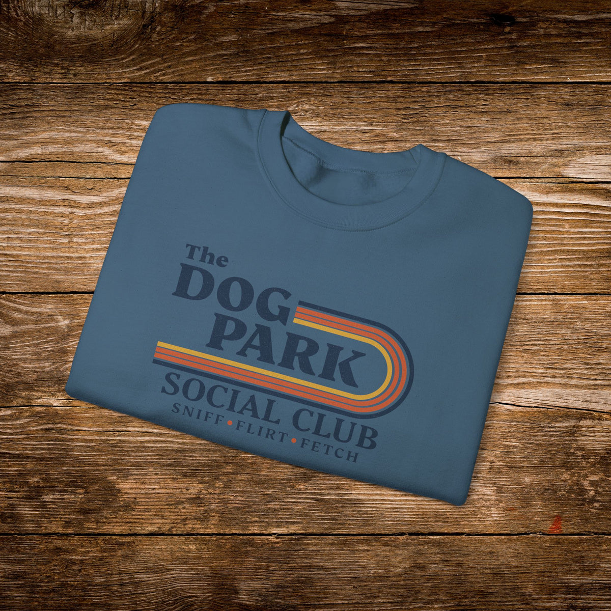 A folded slate blue t-shirt with a retro-style graphic reading 'The Dog Park Social Club' and 'Snief * Flirt * Fetch' in dark blue text, styled on a rustic wooden plank background.