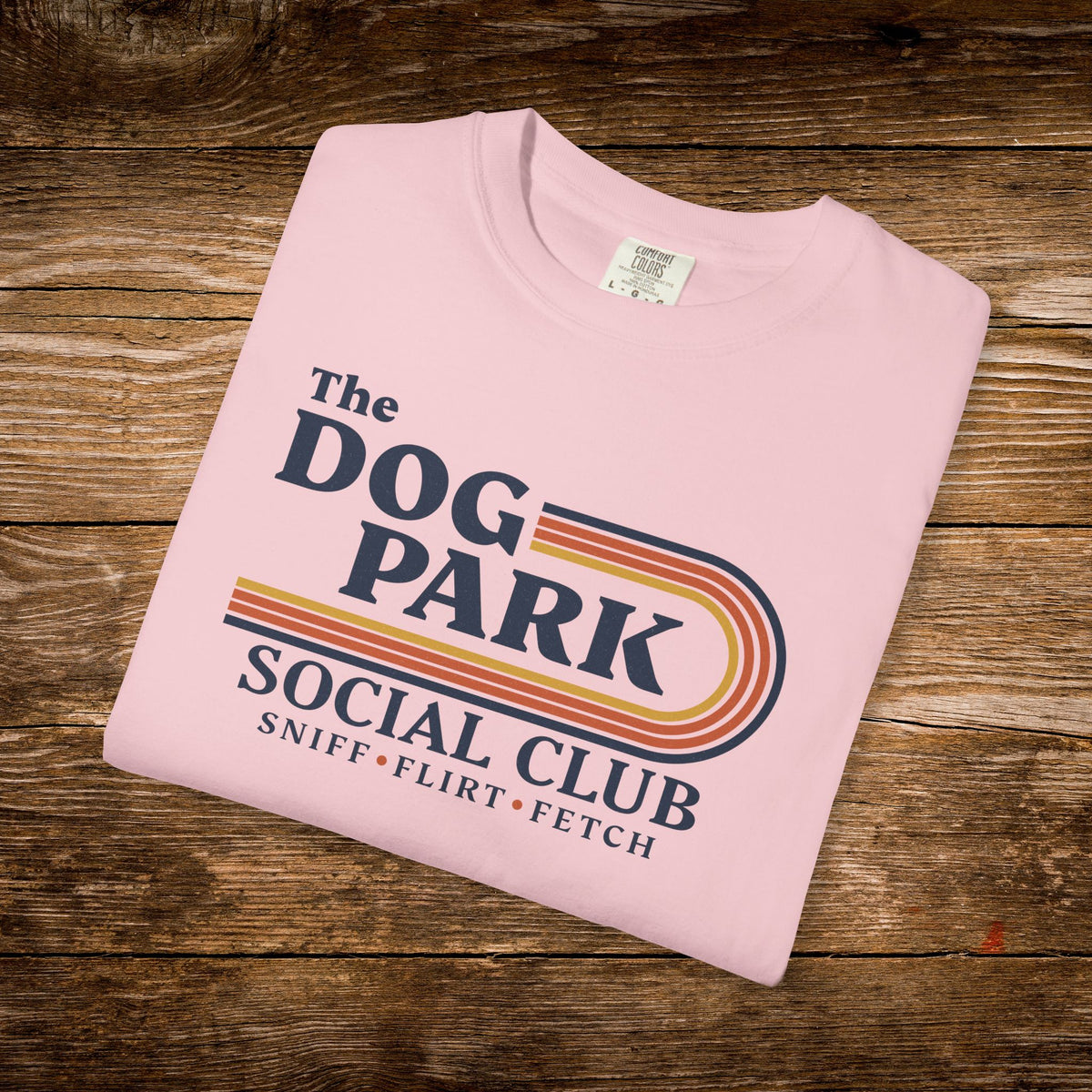A folded light pink t-shirt with a retro-style graphic reading 'The DOG PARK SOCIAL CLUB' and 'SNIFF • FLIRT • FETCH' in navy and orange-yellow stripes, displayed on a rustic wooden surface.