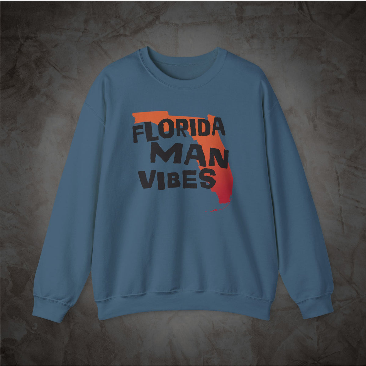 A dusty blue crewneck sweatshirt featuring a graphic print of the state outline of Florida with the text 'FLORIDA MAN VIBES' in black, styled with orange and red paint splatters, displayed on a textured dark gray background.