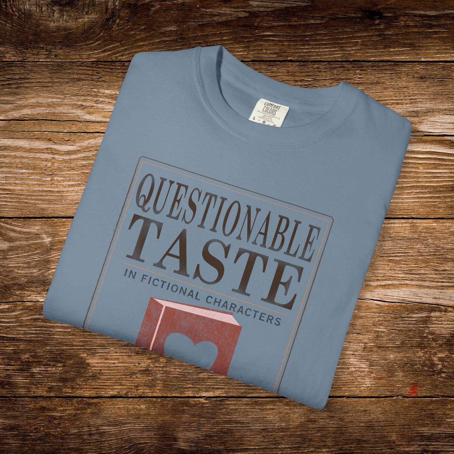 Folded blue t-shirt with 'Questionable Taste' graphic on a wooden surface