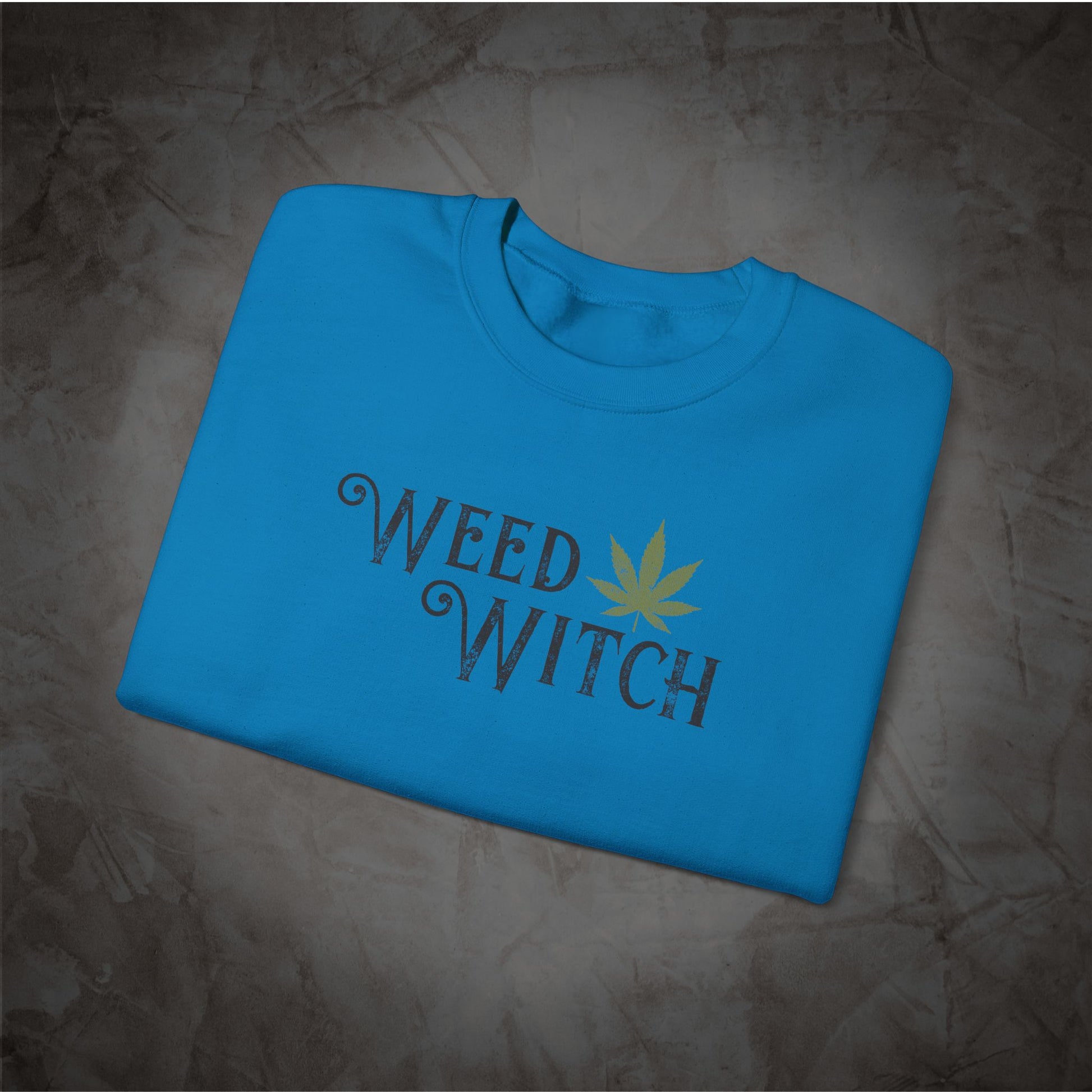 A folded bright blue t-shirt with a graphic print reading 'WEED WITCH' in a dark gray gothic font, featuring a yellow cannabis leaf replacing the letter 'O', displayed on a textured gray background.