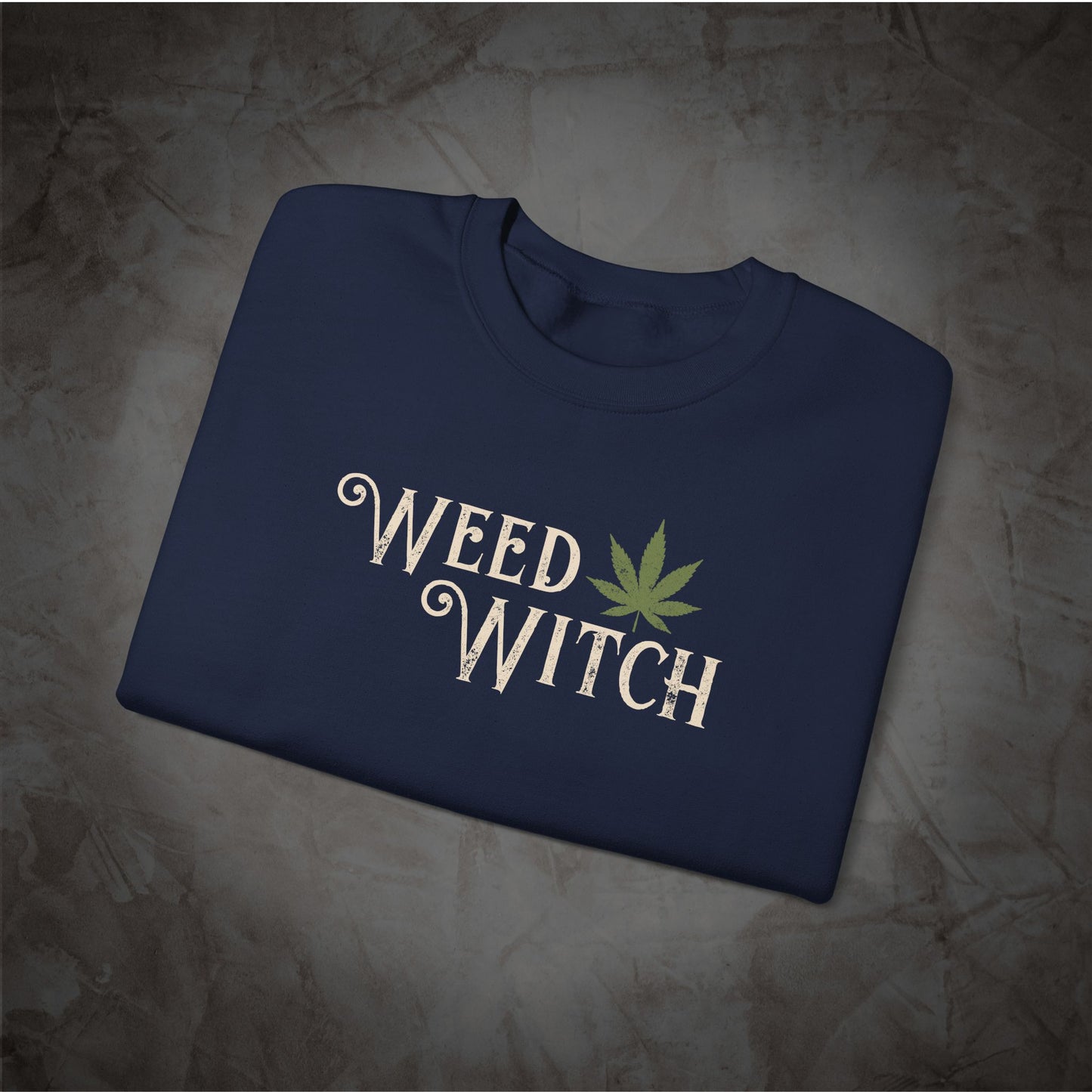 A navy blue folded t-shirt with 'WEED WITCH' printed in white script and a green cannabis leaf graphic, displayed on a textured gray background.