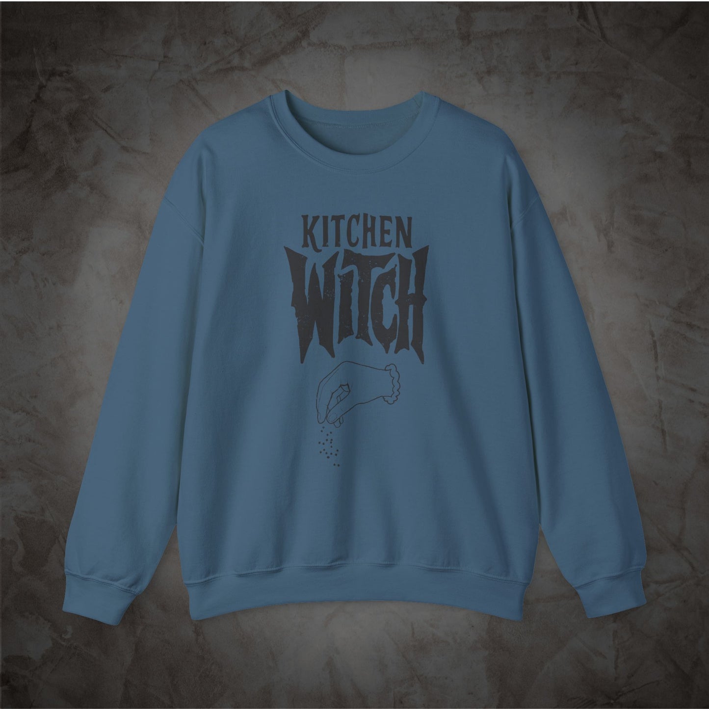 A dusty blue crewneck sweatshirt featuring a dark gray graphic print that reads 'KITCHEN WITCH' with a hand sprinkling salt, displayed against a textured dark gray background.