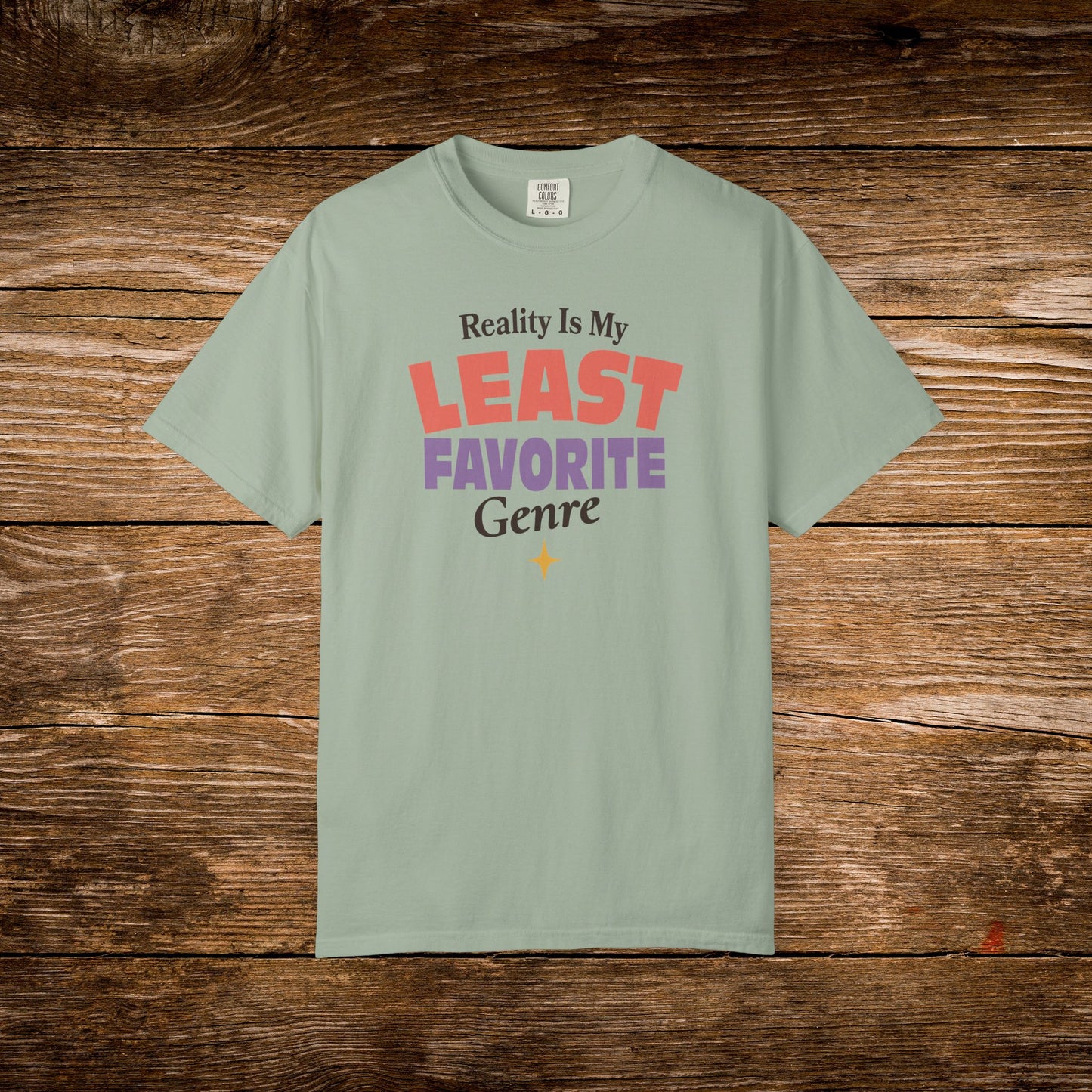 A sage green short-sleeve t-shirt with a colorful graphic text print reading 'Reality Is My LEAST FAVORITE Genre' on a rustic brown wooden background.