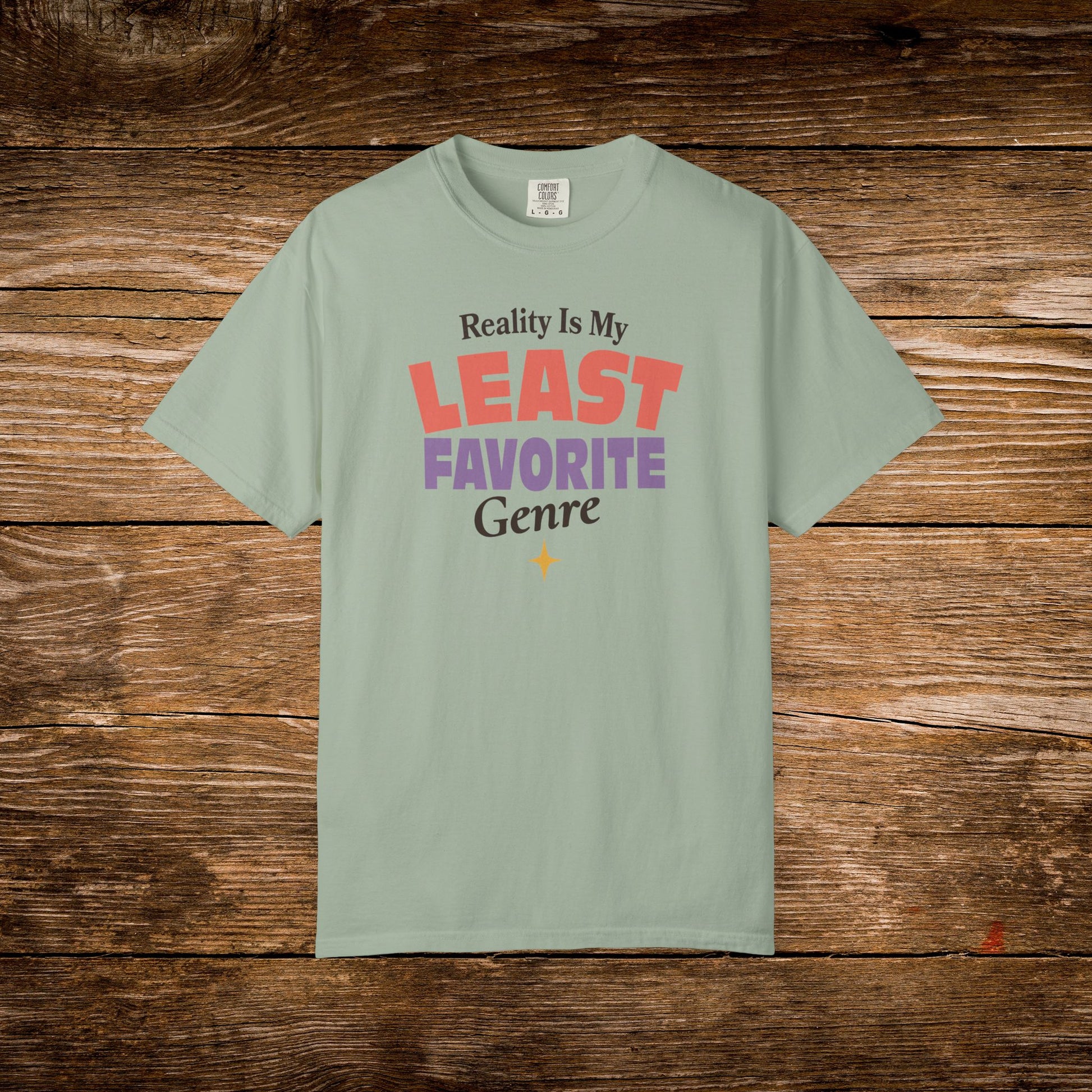 A sage green short-sleeve t-shirt with a colorful graphic text print reading 'Reality Is My LEAST FAVORITE Genre' on a rustic brown wooden background.