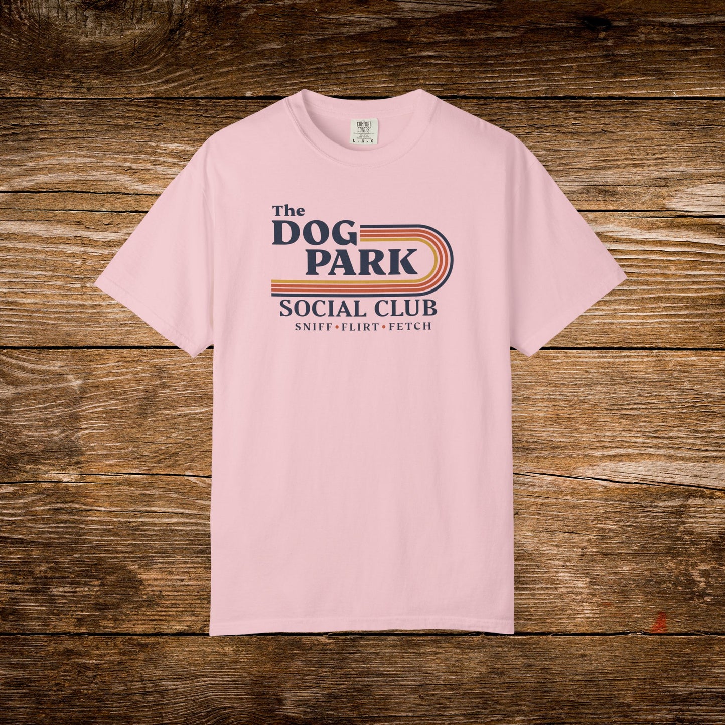 A light pink short-sleeve t-shirt with a retro-style graphic reading 'The DOG PARK SOCIAL CLUB' and 'SNIFF - FLEBIT - FETCH' in navy and orange, laid flat on a rustic brown wooden background. The design text reads "Sniff, Flirt, Fetch."