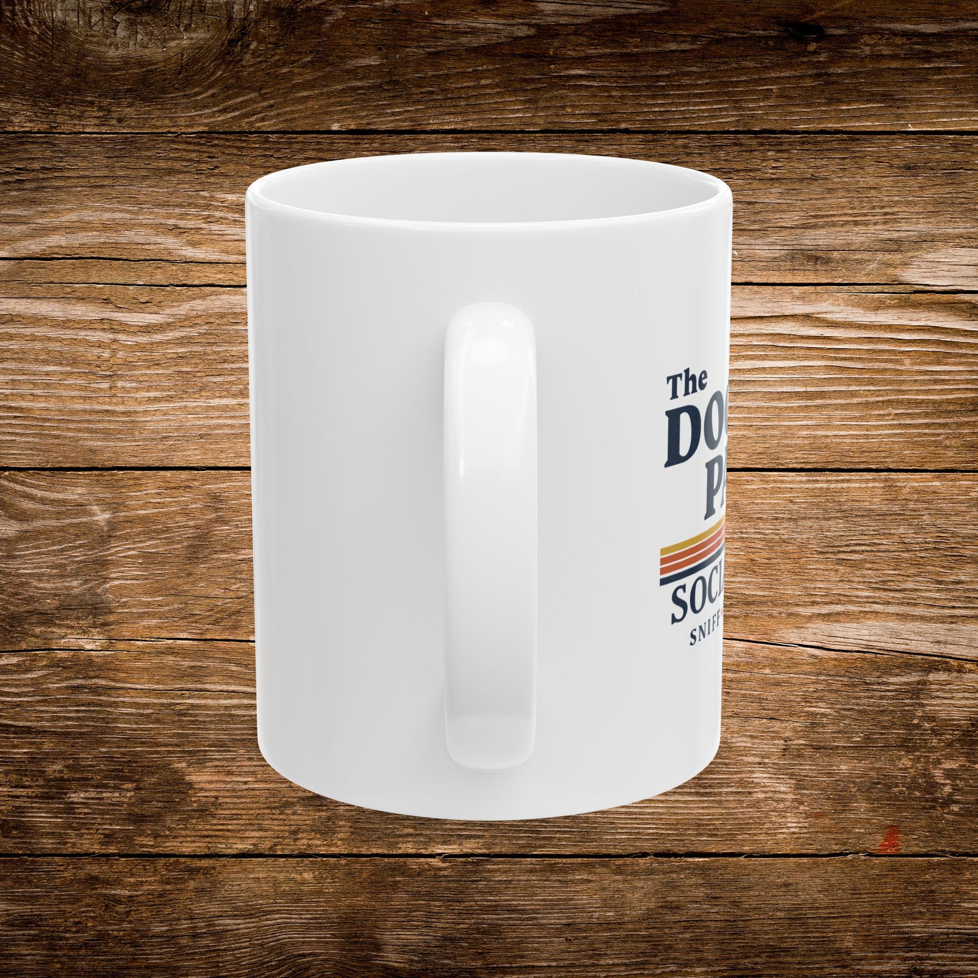 A white ceramic coffee mug with a printed graphic on the side, displayed on a rustic brown wooden plank background. The graphic features the text 'The DOG PUP' and 'SOCIAL' in dark blue lettering with a retro orange and yellow stripe design. The design text reads "Sniff, Flirt, Fetch."