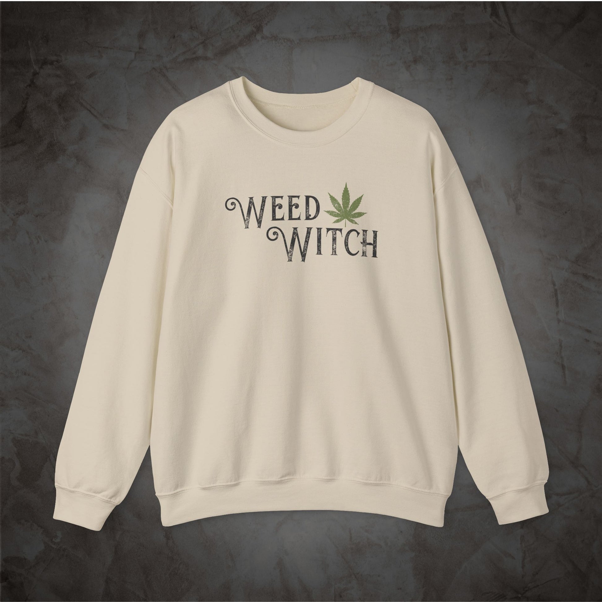 An off-white crewneck sweatshirt featuring the words 'WEED WITCH' in a dark gray gothic font with a green cannabis leaf graphic, displayed against a dark textured background.
