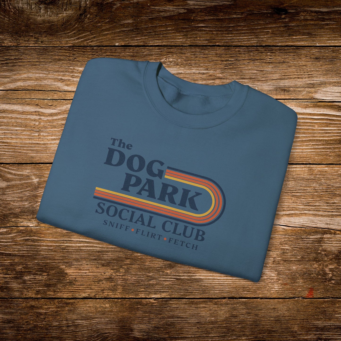 A folded slate blue t-shirt with a retro-style graphic reading 'The Dog Park Social Club' and 'Sniff, Flirt, Fetch' in yellow, orange, and red stripes, displayed on a rustic wooden plank background.