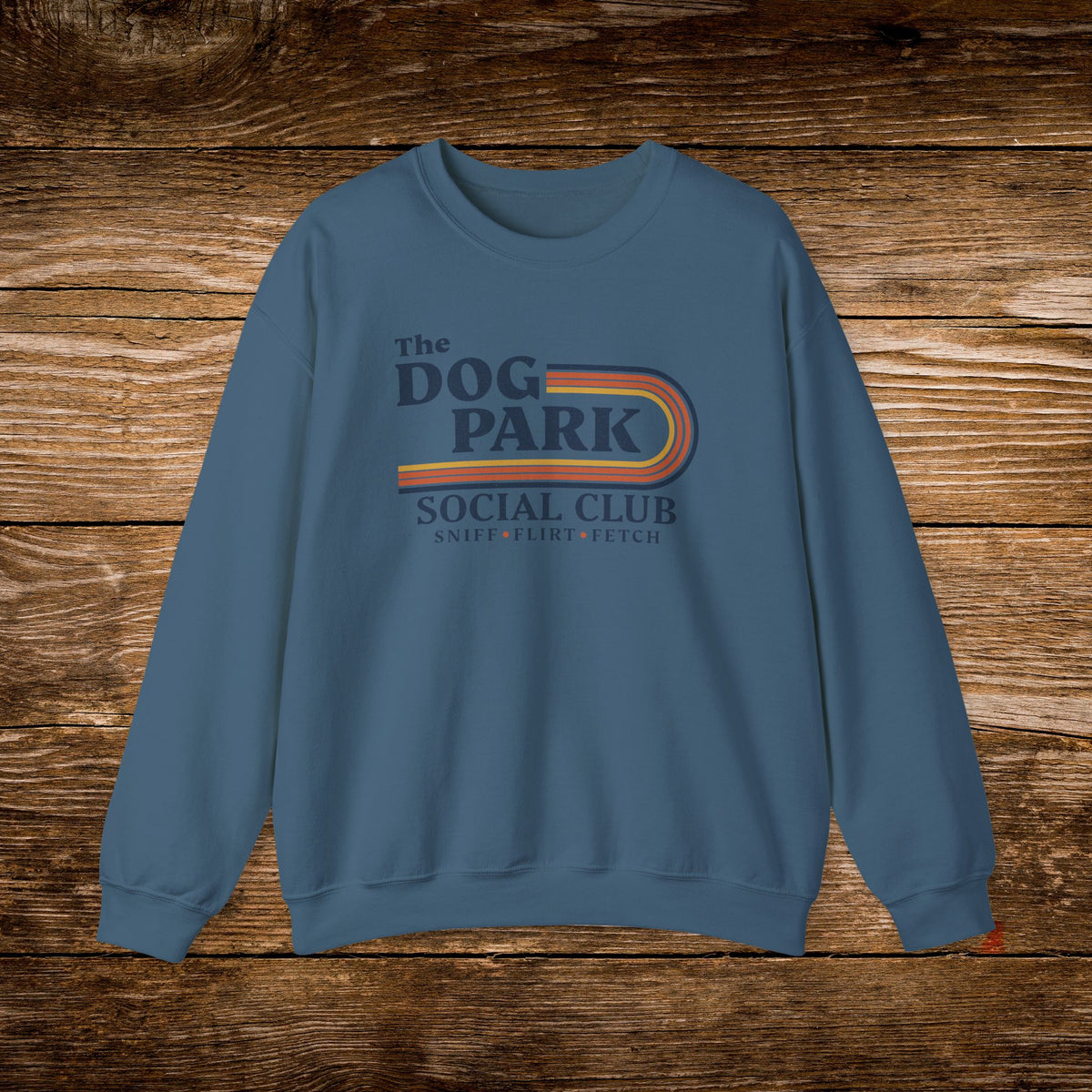 A slate blue crewneck sweatshirt featuring a retro-style graphic that reads 'The DOG PARK SOCIAL CLUB' with 'SNIPER, PLOYER, FETCH' underneath, displayed flat against a rustic brown wooden plank background.