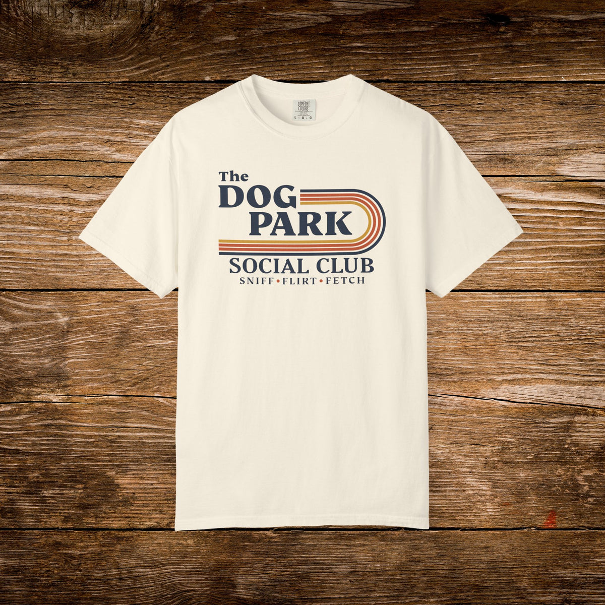 An off-white short-sleeve t-shirt with a retro-style graphic reading 'The DOG PARK SOCIAL CLUB' and 'SNIFF - FLIRT - FETCH' in navy and orange, laid flat on a rustic brown wooden background.