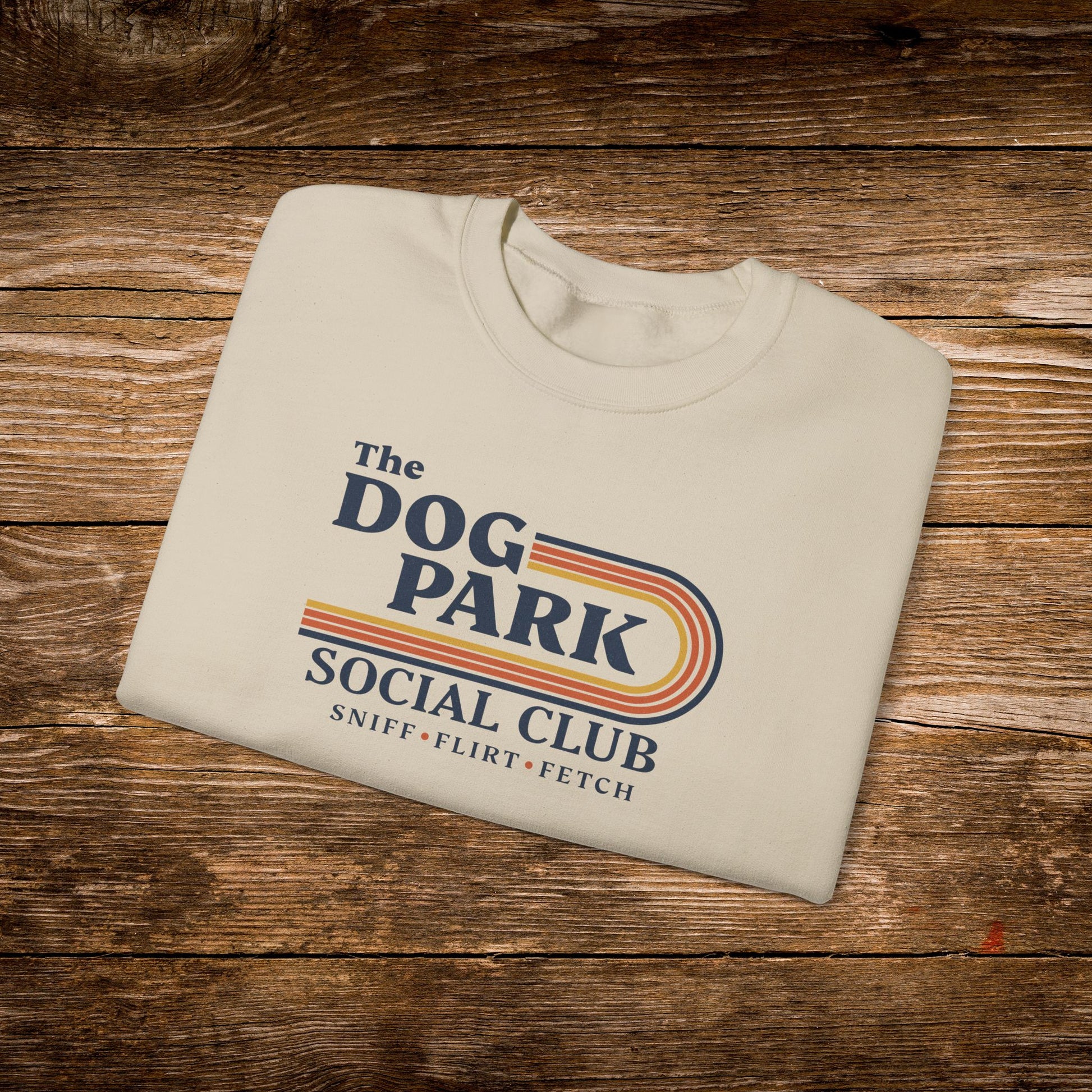 A folded cream-colored t-shirt with a retro-style graphic reading 'The Dog Park Social Club' and 'Sniff • Flirt • Fetch' in purple and pink/yellow stripes, displayed on a rustic wooden background. The design text reads "Sniff, Flirt, Fetch."