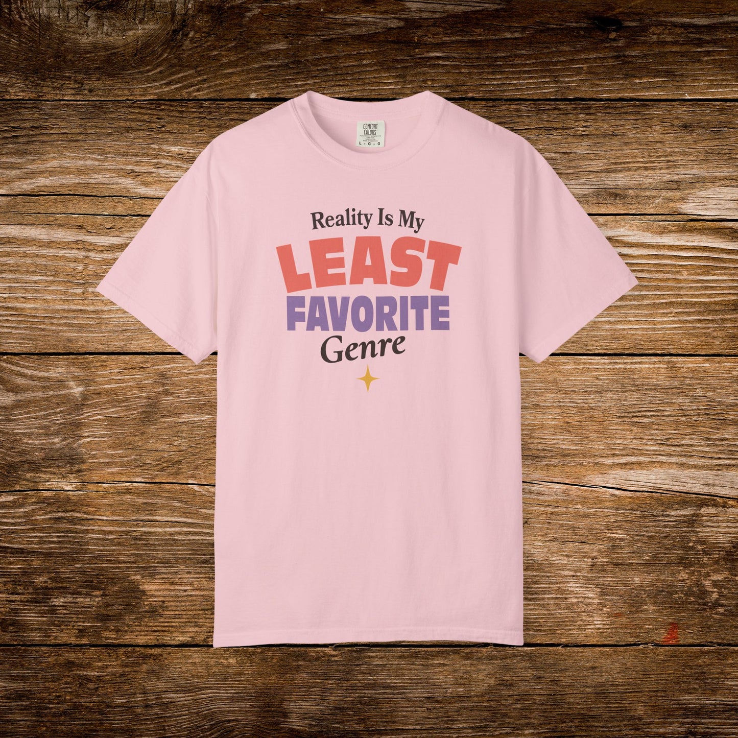 A light pink short-sleeve t-shirt with a graphic text print reading 'Reality Is My LEAST FAVORITE Genre' in red, purple, and black lettering, displayed flat against a rustic brown wooden plank background.