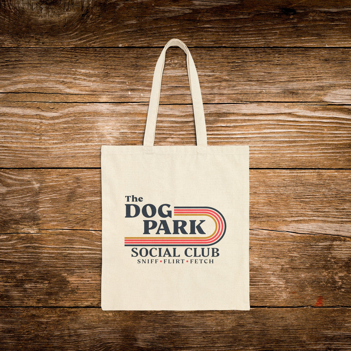 A cream canvas tote bag with 'The Dog Park Social Club' printed on the front, displayed flat against a rustic brown wooden plank background.