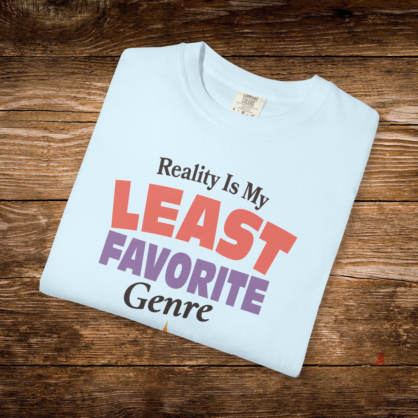 A folded light blue t-shirt with a humorous graphic reading 'Reality Is My LEAST FAVORITE Genre' in red, purple, and black text, displayed on a rustic brown wooden background.