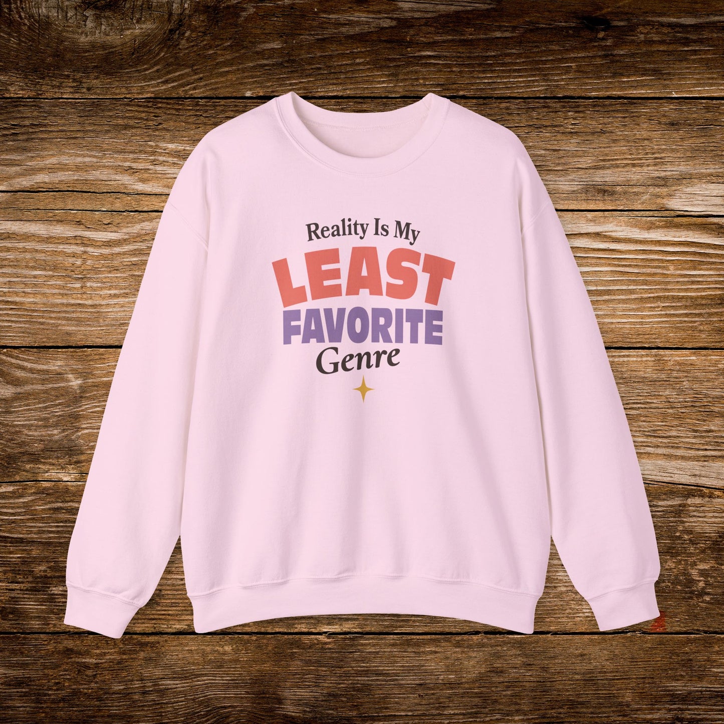 A light pink crewneck sweatshirt with a graphic text print reading 'Reality Is My LEAST FAVORITE Genre' in red, purple, and black lettering, displayed flat on a rustic brown wooden background.