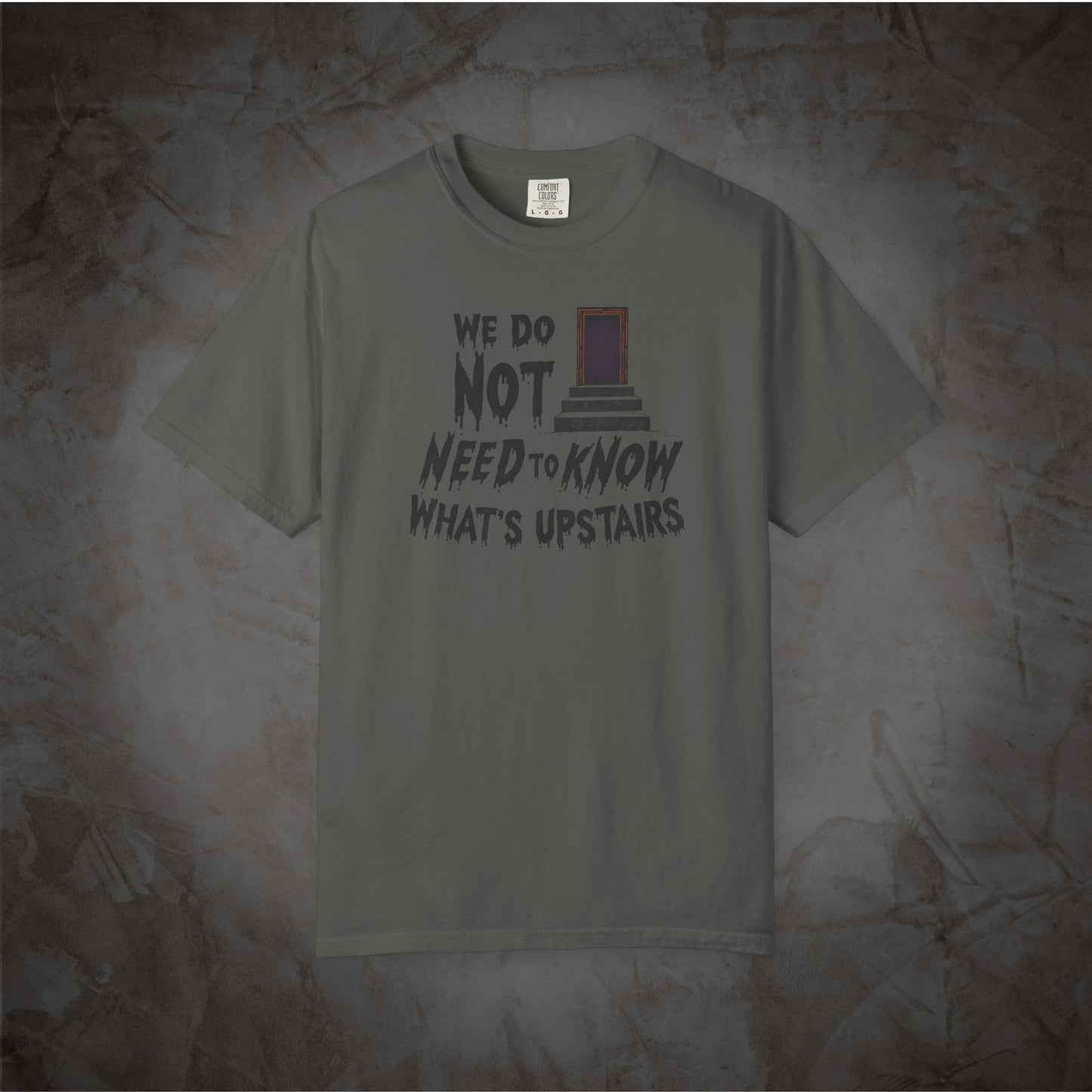 A gray short-sleeve t-shirt with a graphic print on the front that reads 'WE DO NOT NEED TO KNOW WHAT'S UPSTAIRS' alongside a small purple and black door illustration, displayed against a textured gray background.
