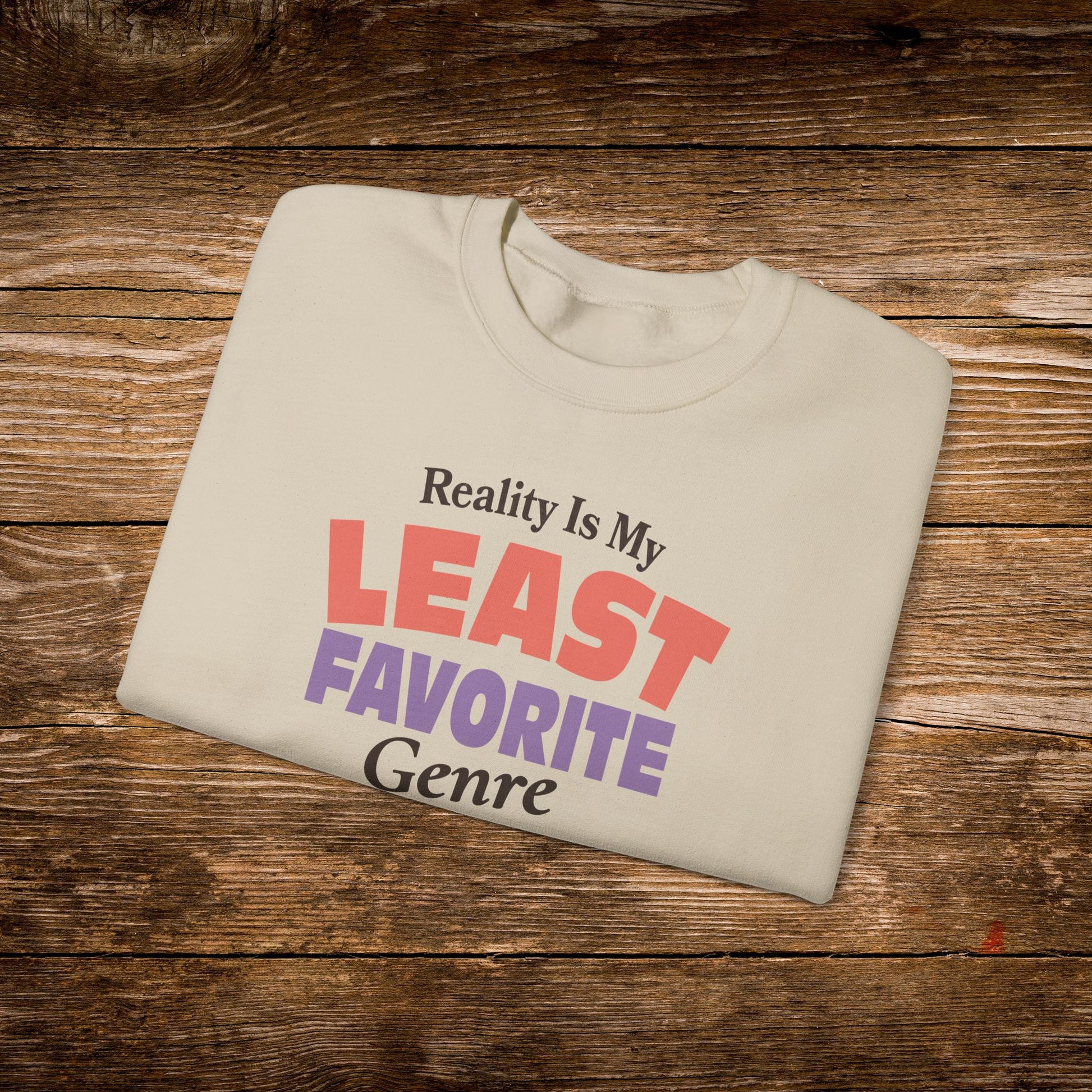 A folded cream-colored t-shirt with the text 'Reality Is My LEAST FAVORITE Genre' printed on the front, displayed on a rustic wooden background.