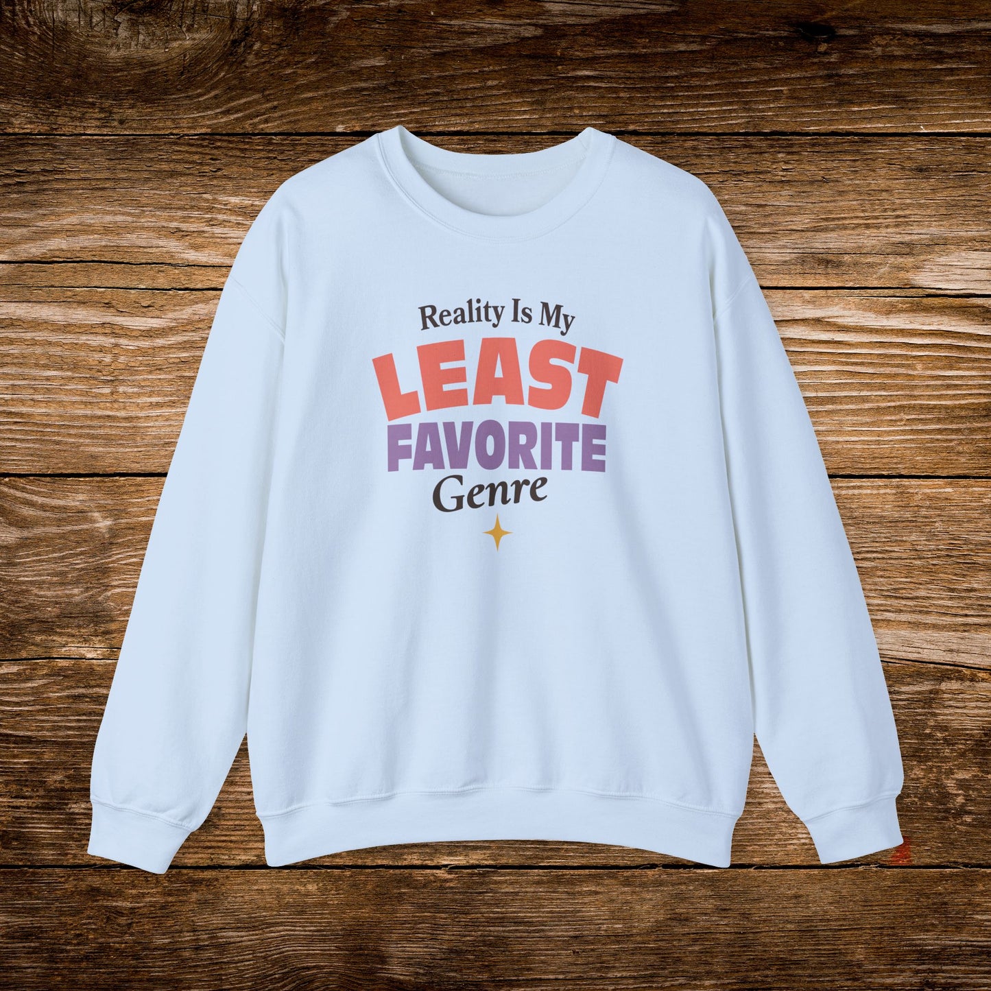 A light blue crewneck sweatshirt with a colorful graphic text print reading 'Reality Is My LEAST FAVORITE Genre' on a rustic dark wood background.