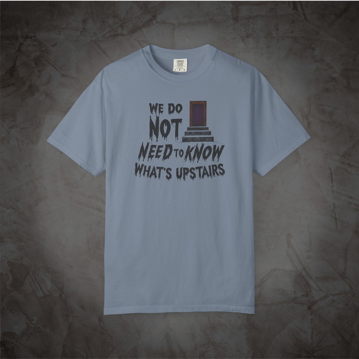 A light blue crewneck t-shirt with a graphic print on the front that reads 'WE DO NOT NEED TO KNOW WHAT'S UPSTAIRS' alongside a drawing of the TARDIS, displayed against a textured dark gray background.